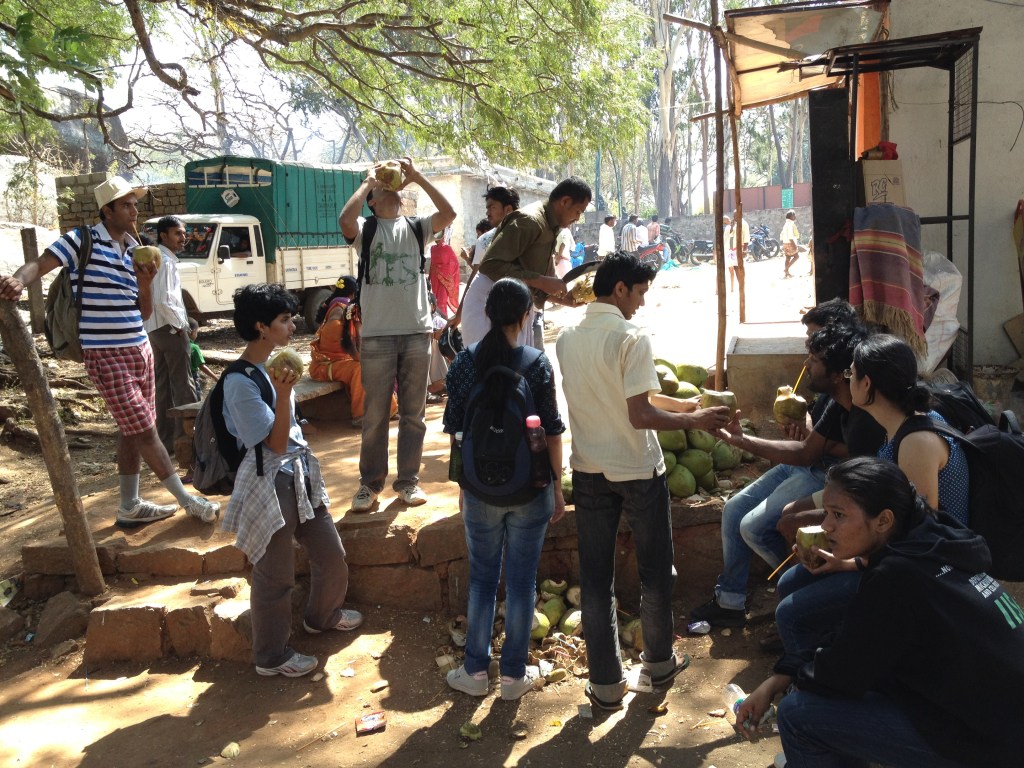 Group photo from a lab outing showing Dr. Amit Agrawal, Prof. Vishwesha (Vishu) Guttal, and Dr. Jaideep Joshi with their team from IISc drinking coconut juice during a hike at Savandurga Hills.